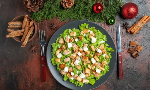 Top view caesar salad on oval plate fork knife bowl with dried crust xmas tree toys cinnamon sticks on dark red background
