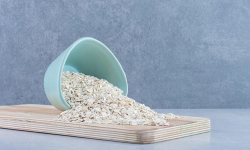 Rolled oats spilling from a small bowl onto a wooden board against a gray marble background