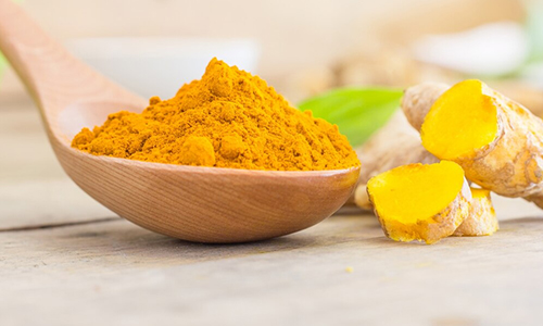 Wooden spoon with turmeric powder next to fresh turmeric root on a wooden table.