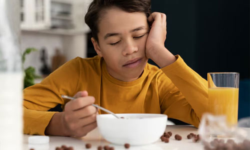 A tired young boy sitting at a breakfast table, resting their head on one hand with a bowl of cereal and a glass of juice in front of them, showing the effects of skipping or delaying breakfast.