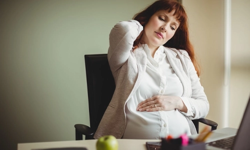 Pregnant businesswoman sitting in a chair holding her back, used to illustrate the risk of Deep Vein Thrombosis (DVT) during pregnancy.