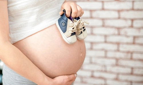 Pregnant woman holding small baby shoes against her belly, representing the journey of gestational surrogacy.