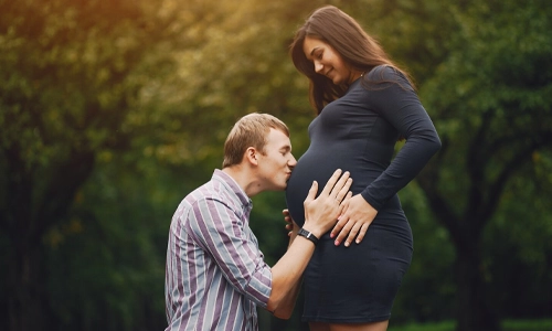 A happy couple kissing his pregnant wife belly in a park with green trees in the background.