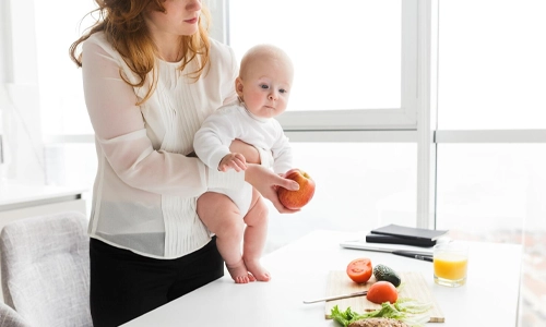 Mother holding her baby while preparing a healthy meal in the kitchen to support milk production and healing.