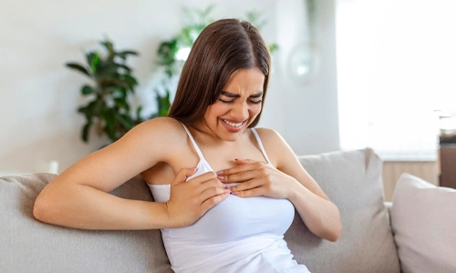 Woman sitting at home with a pained expression holding her chest, representing symptoms of postpartum mastitis.