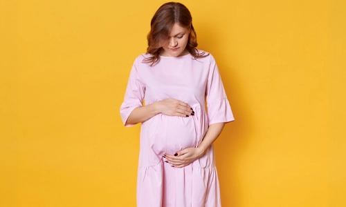 Pregnant woman in pink dress gently holding her belly against a yellow background