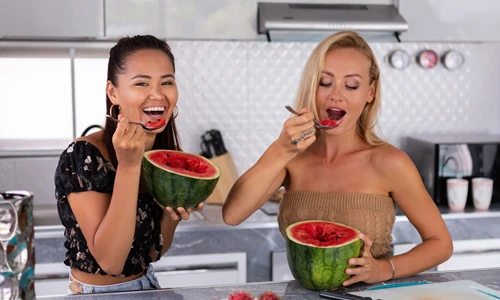 Two women eating half sliced watermelon with spoon at home